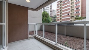 Un balcon doté d’une balustrade en verre, d’un sol en béton et offrant une vue sur un immeuble à plusieurs étages, lui-même pourvu de balcons.