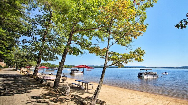 Une plage au bord du lac, avec des tables de pique-nique et des parasols, des bateaux sur l’eau et des arbres le long de la rive.