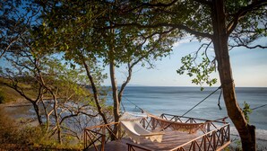 Un hamac sur une terrasse en bois donnant sur une plage bordée d’arbres et sur l’océan.