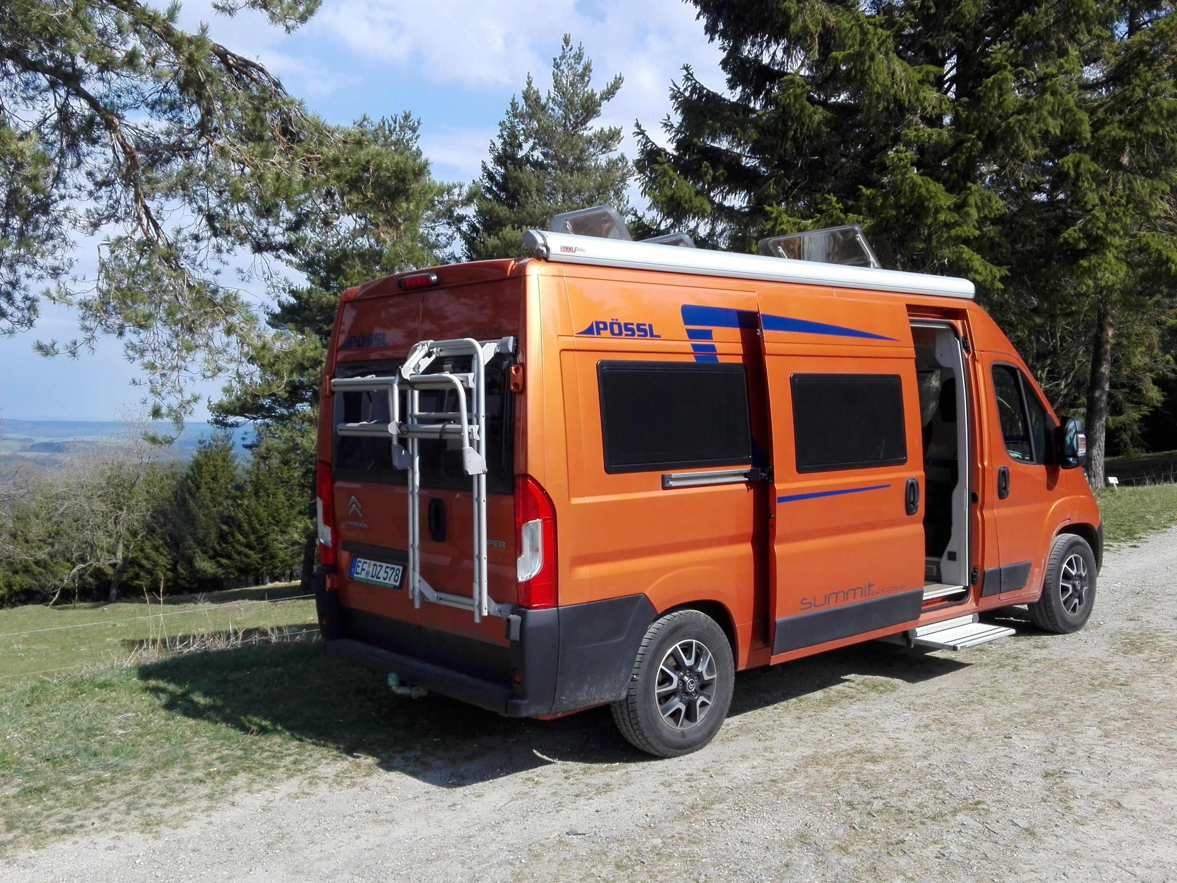 Ein orangefarbener Van mit geöffneter Seitentür, geparkt auf einem Schotterweg mit Bäumen und Blick ins Freie.