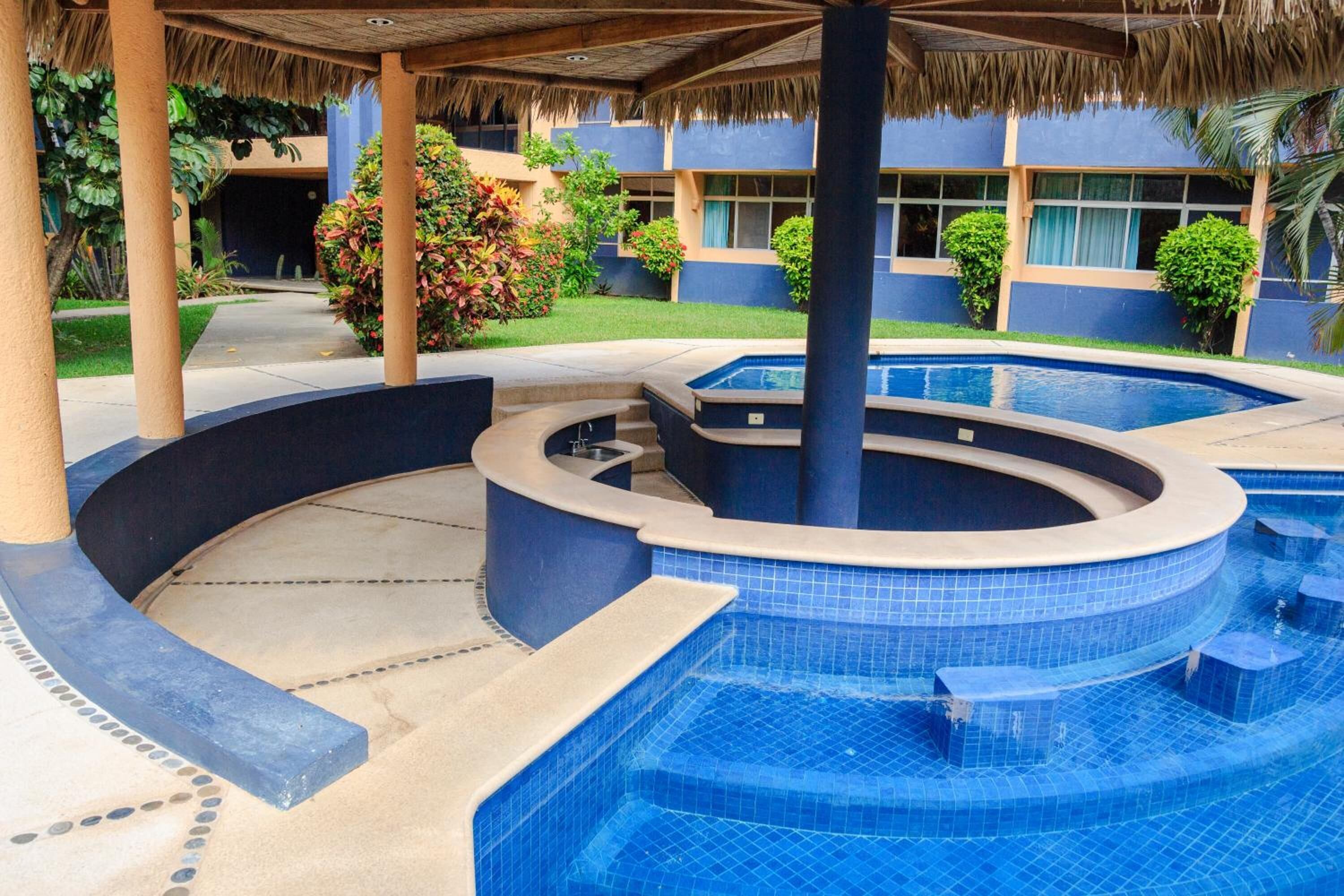 A pool area with a circular hot tub, a covered seating area, and a building with a blue facade in the background.