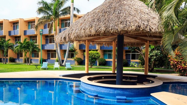 A swimming pool with a thatched roof structure, surrounded by palm trees and a building with balconies. - Hotel Calli (Tehuantepec)