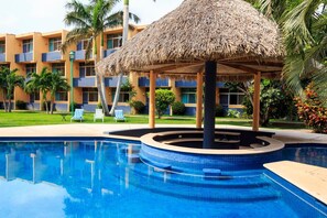 A swimming pool with a thatched roof structure, surrounded by palm trees and a building with balconies. - Hotel Calli (Tehuantepec)