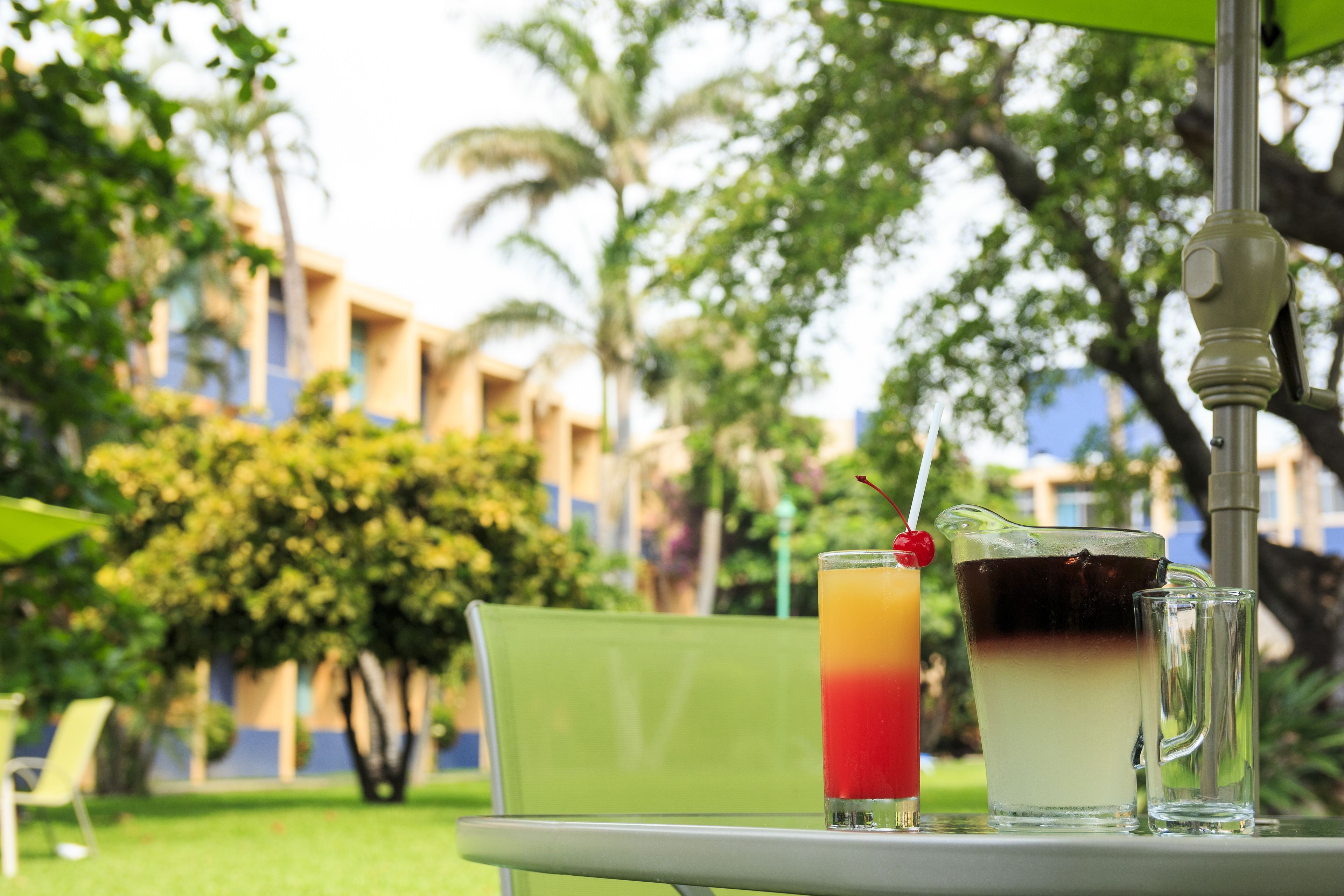A table with two drinks, one with a layered appearance and a cherry on top, and a clear glass next to it. The background features a building with blue and beige walls and lush greenery.