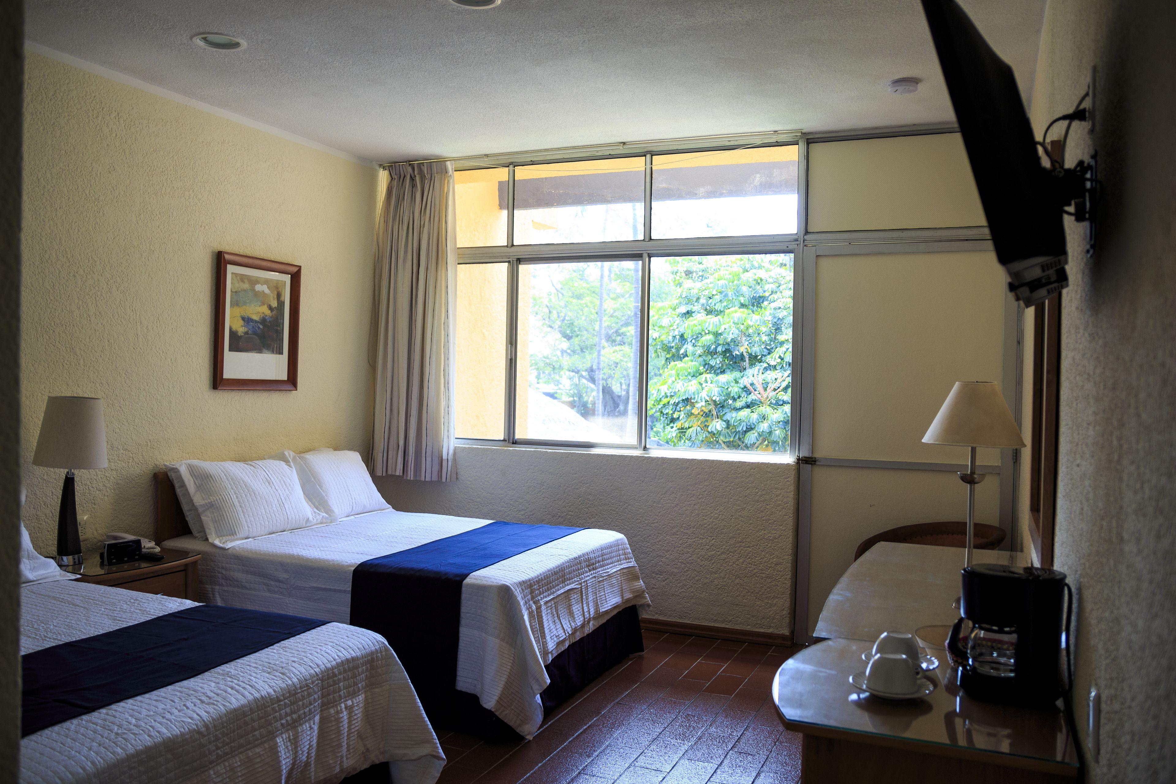 A hotel room with two beds, a window with a view of trees, a desk with a lamp, and a coffee maker.
