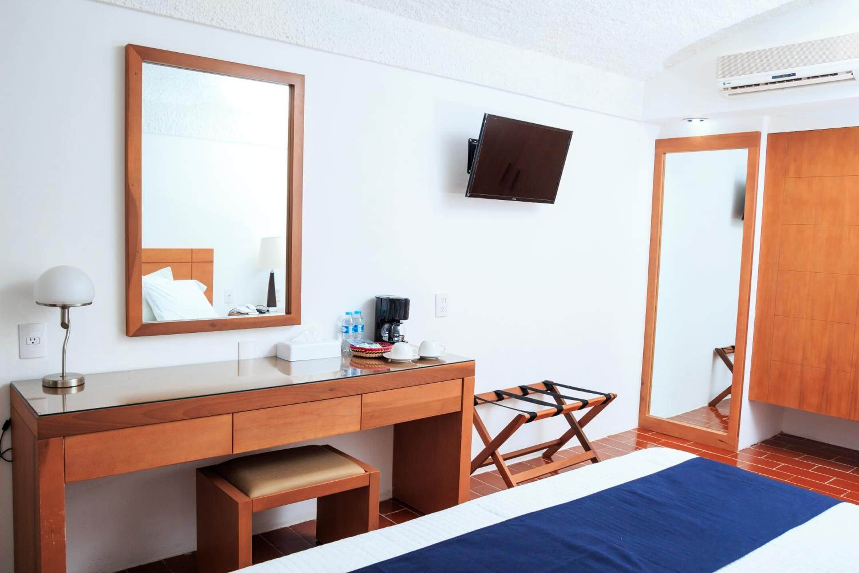 A hotel room with a wooden desk, a bed with a blue bedspread, a mounted TV, and a wardrobe.