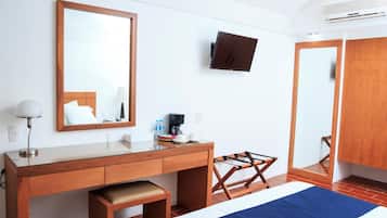 A hotel room with a wooden desk, a bed with a blue bedspread, a mounted TV, and a wardrobe.