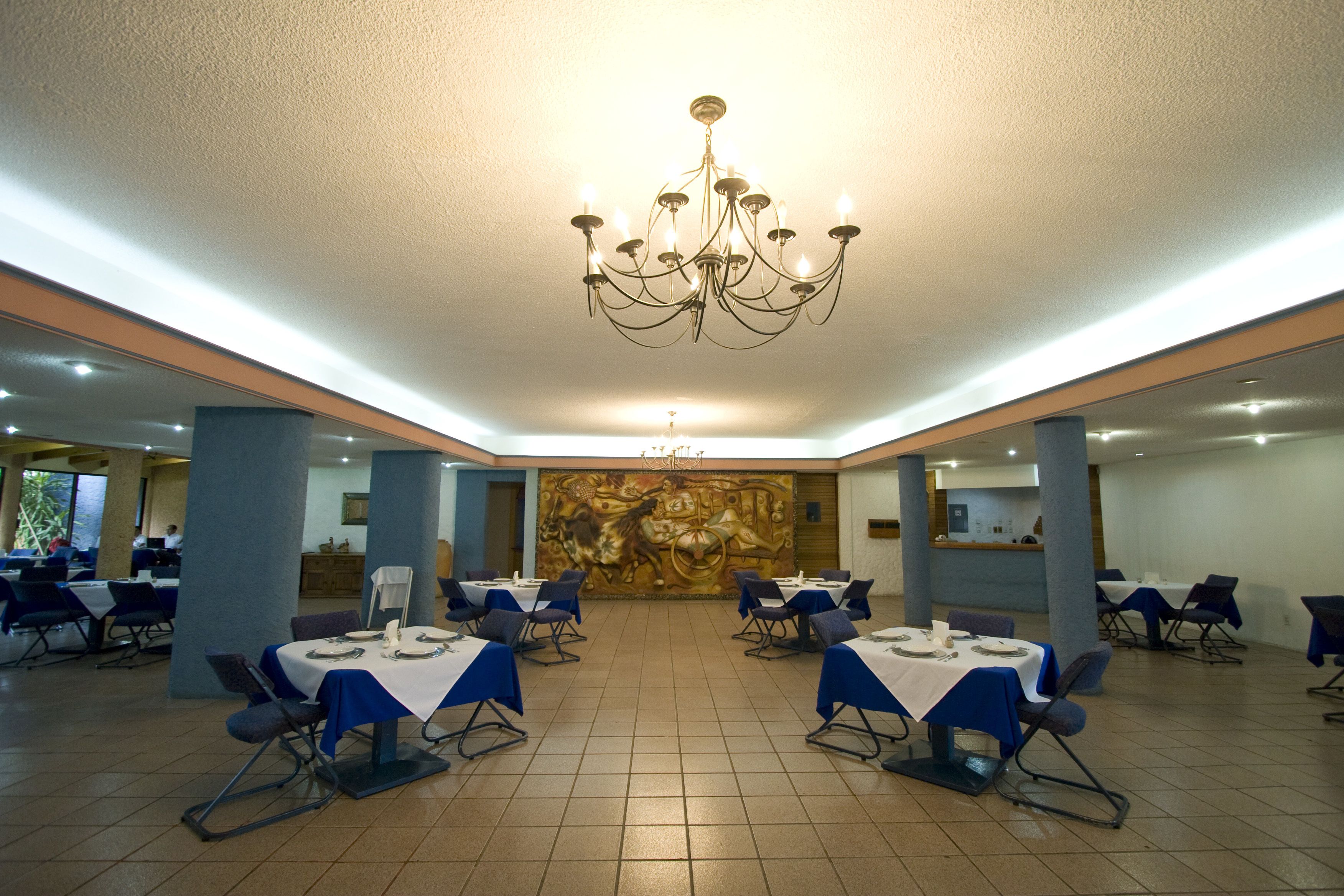 Un comedor amplio con mesas dispuestas para el servicio, una lámpara de araña grande y un mural en la pared de fondo.