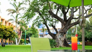 An outdoor table with a green umbrella, a glass of red drink with a cherry, and a large tree in the background. - Hotel Calli (Tehuantepec)