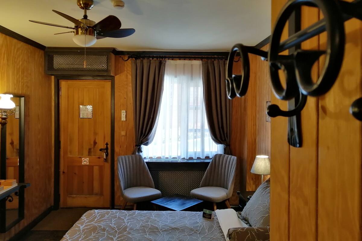 A hotel room with a bed, two chairs, a desk, a ceiling fan, and a window with curtains.