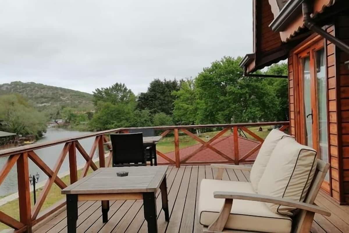 A wooden deck with a table and a chair, overlooking a river and hills.