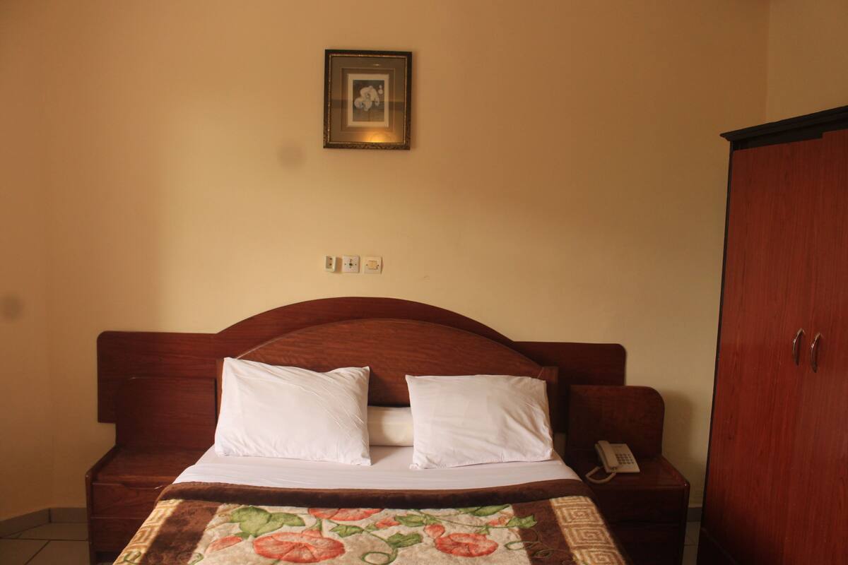 A bedroom with a wooden bed, white pillows, a floral bedspread, a bedside telephone, and a wardrobe.