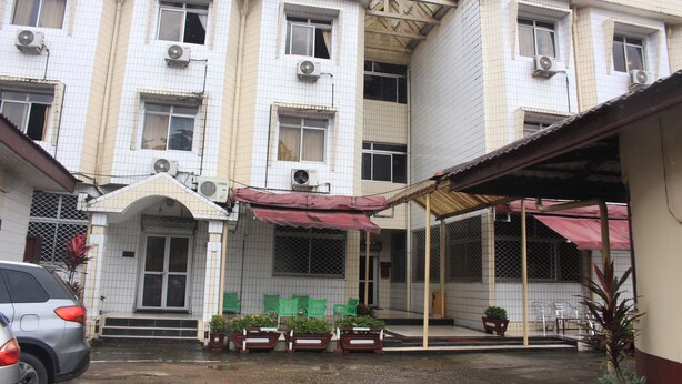 A multi-story building with tiled exterior walls, multiple air conditioning units, and a covered entrance area with chairs and potted plants. - Hôtel Beau Rivage (Douala)