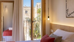 Ein Schlafzimmer mit Balkon, einem Bett mit rot-weißer Bettwäsche und Blick auf ein Steinhaus und Grünflächen.