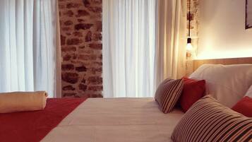 Habitación de hotel con pared de ladrillo, cortinas blancas, cama con almohadas rojas y a rayas, y una lámpara de noche.