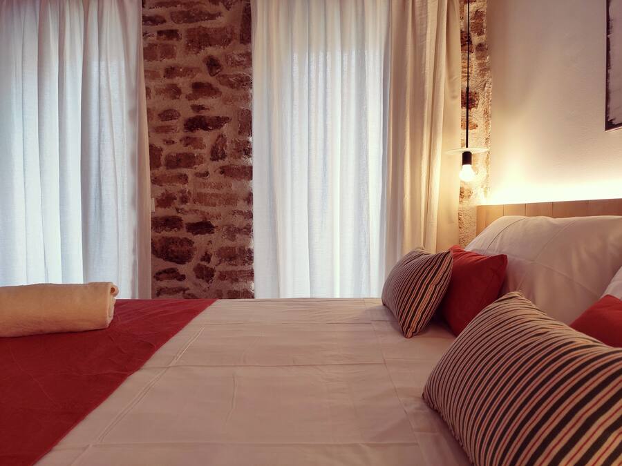 Habitación de hotel con pared de ladrillo, cortinas blancas, cama con almohadas rojas y a rayas, y una lámpara de noche.