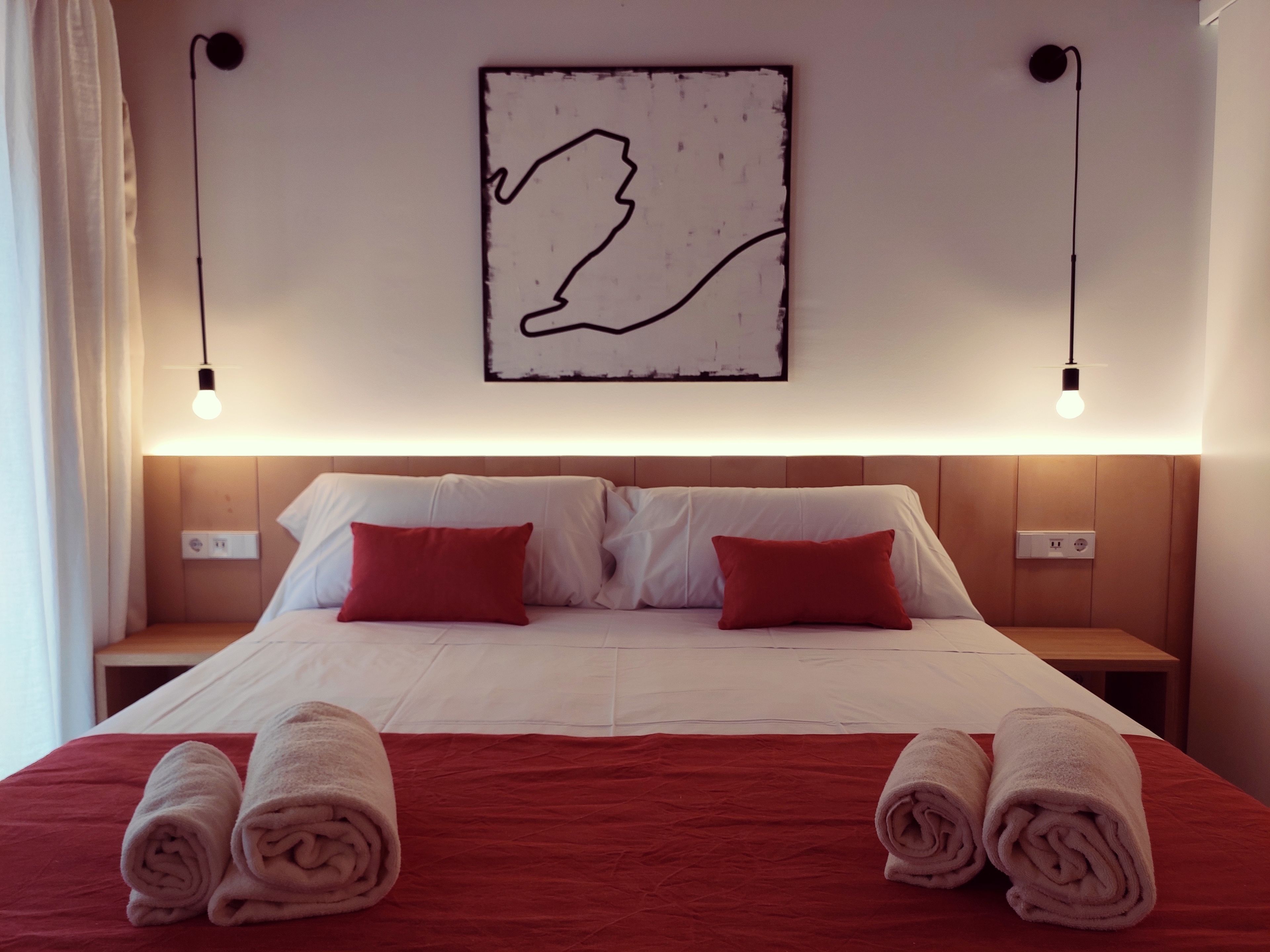 A neatly made bed with red and white bedding, two red pillows, and rolled towels on the side.