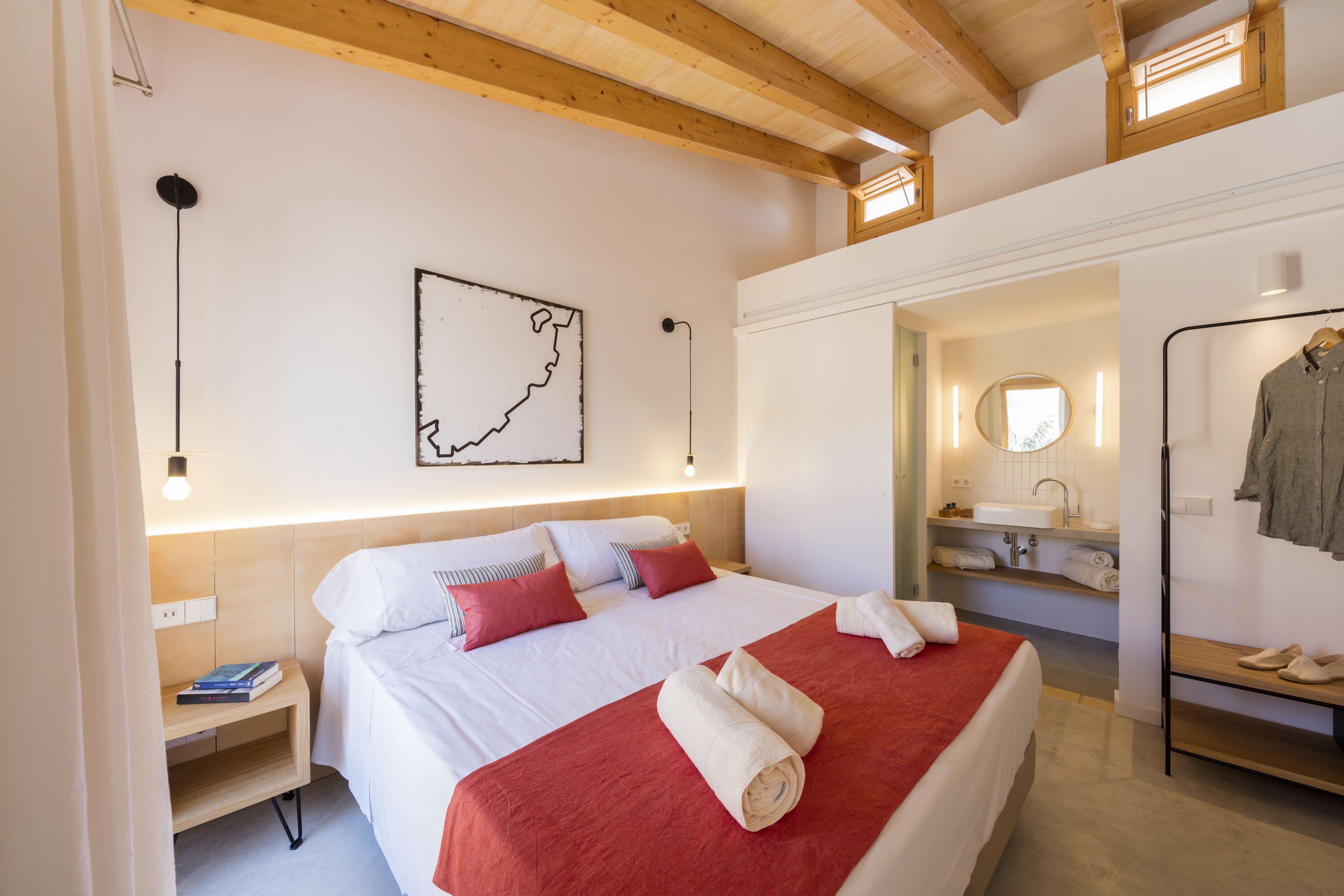 Habitación de hotel con techo de madera, cama con ropa de cama blanca y roja, un mapa en la pared, una mesita de noche con libros, y un baño con espejo y lavamanos.