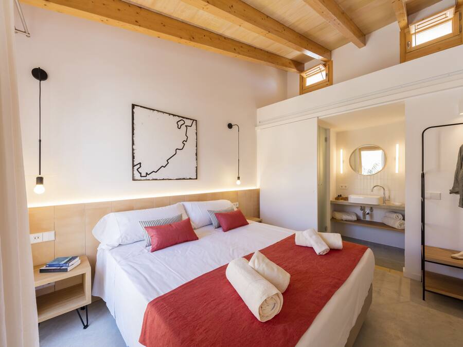 Habitación de hotel con techo de madera, cama con ropa de cama blanca y roja, un mapa en la pared, una mesita de noche con libros, y un baño con espejo y lavamanos.
