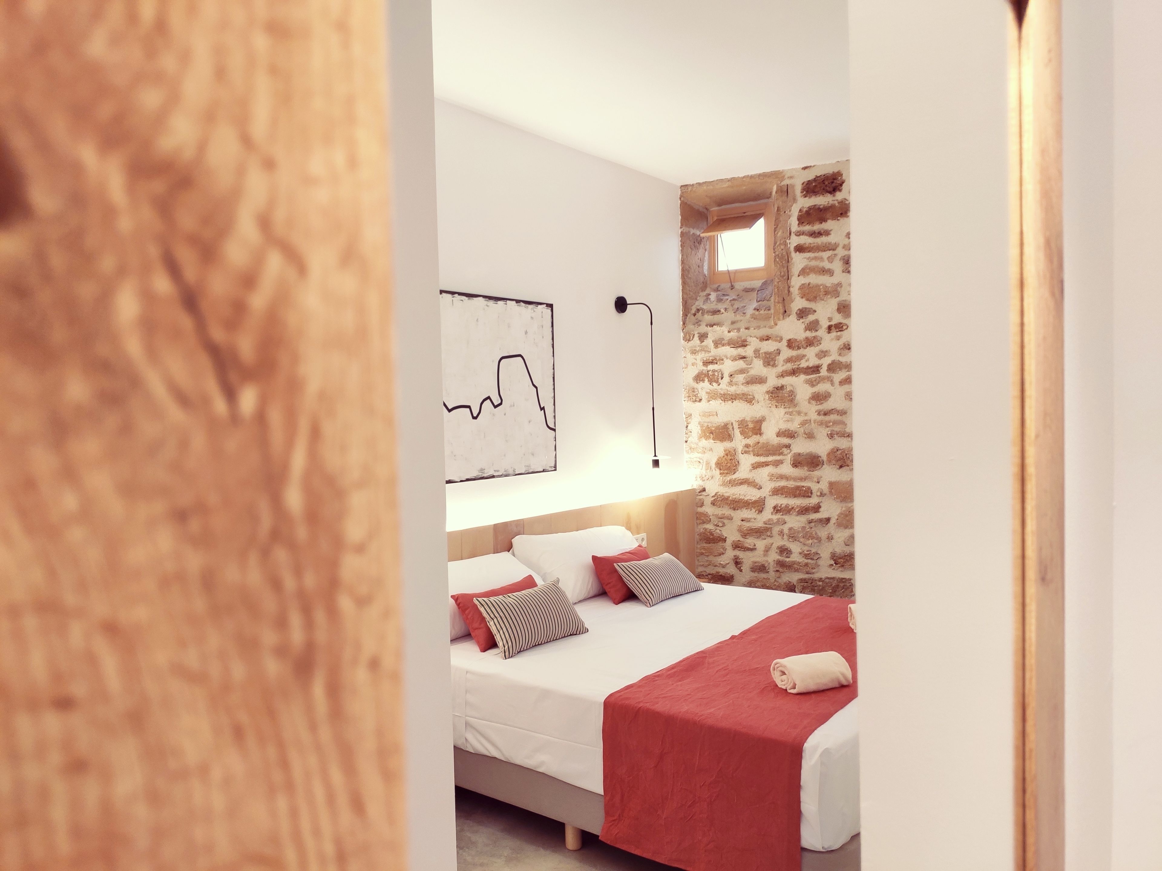 A modern bedroom with a stone wall, a bed with white bedding and red accents, a wall-mounted lamp, and a window.