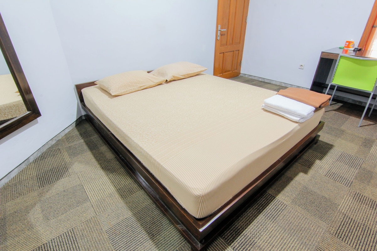 A neatly made bed with a wooden frame and a beige mattress, a wooden door, and a small desk with a chair.
