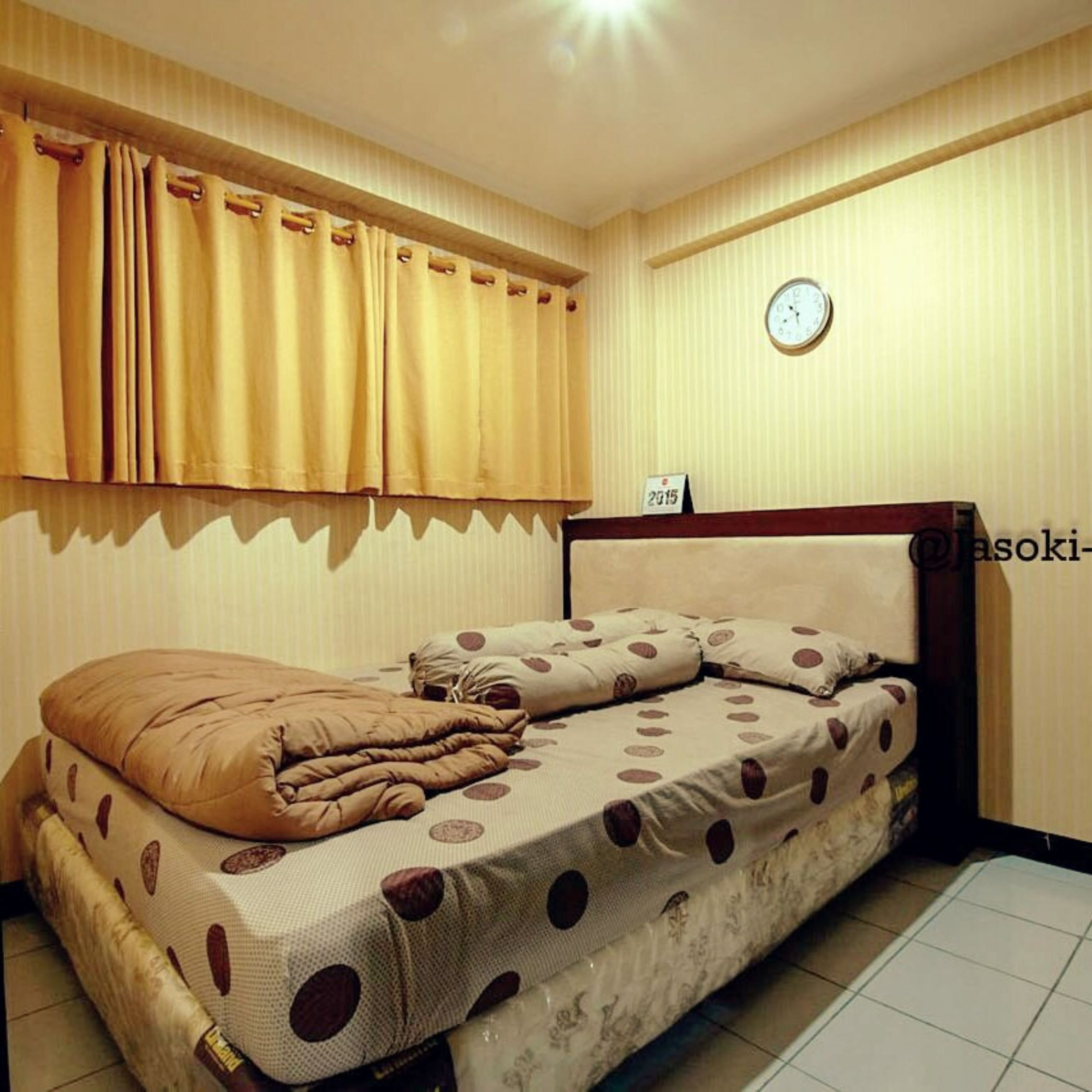 A small, single-bed room with a wooden headboard, a beige bedspread with a polka dot pattern, and a window with yellow curtains.