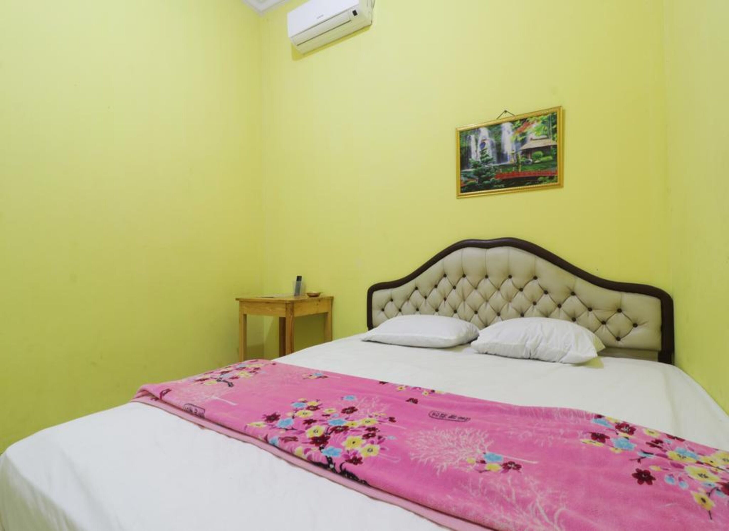 Una habitación con una cama, una mesita de noche de madera, un aire acondicionado de pared y un cuadro enmarcado.