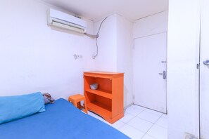 Una habitación con una cama, un pequeño estante naranja, una puerta blanca y un equipo de aire acondicionado.
