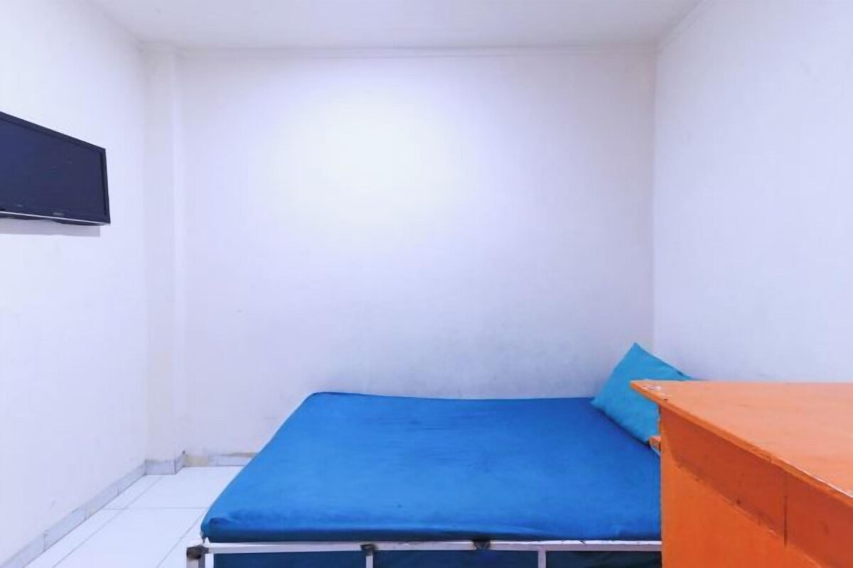 A small, single-bed room with a blue mattress, a wooden nightstand, and a wall-mounted TV.