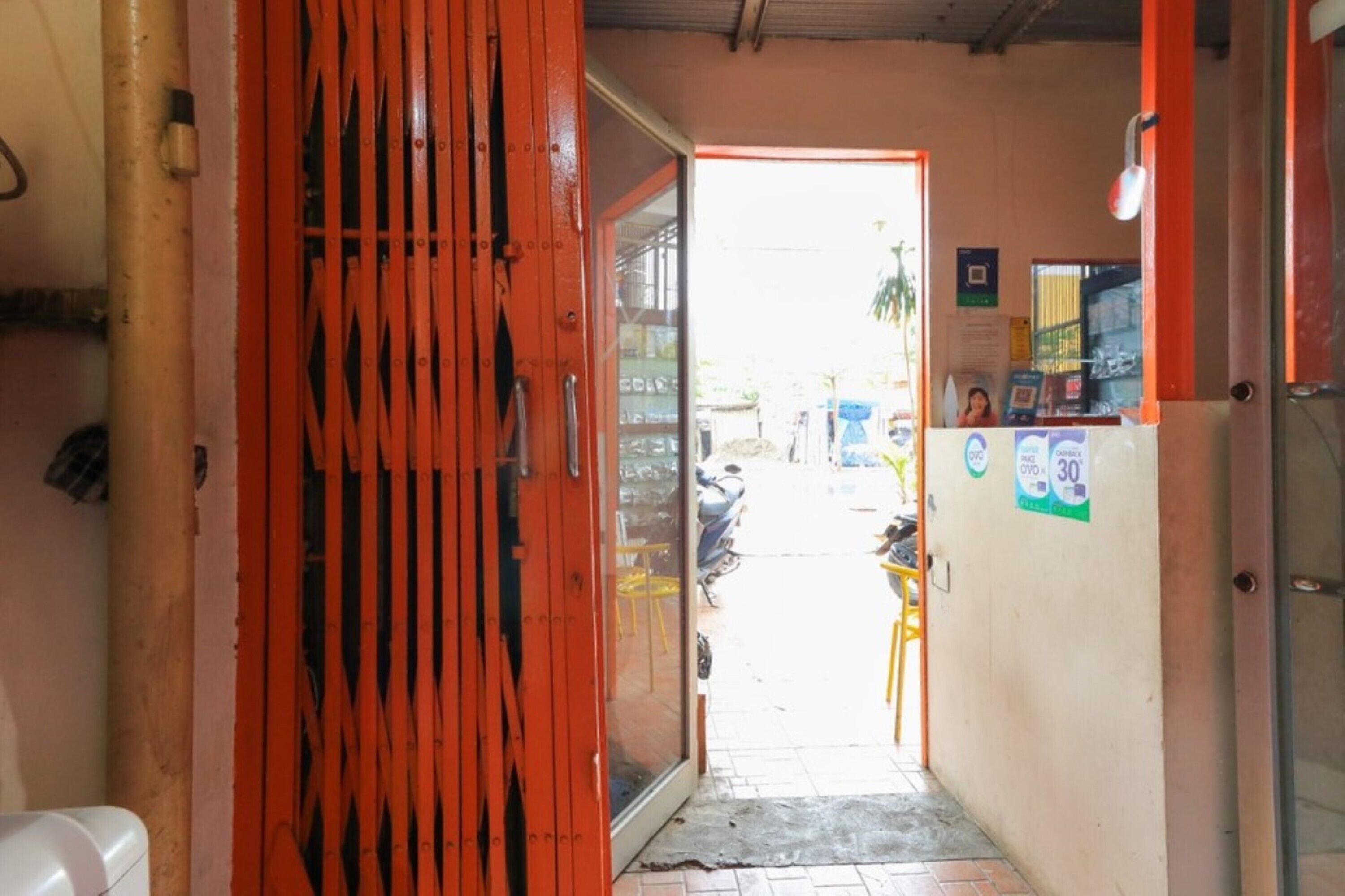 A narrow hallway with a red door leading to a brightly lit area with a scooter and a yellow chair.