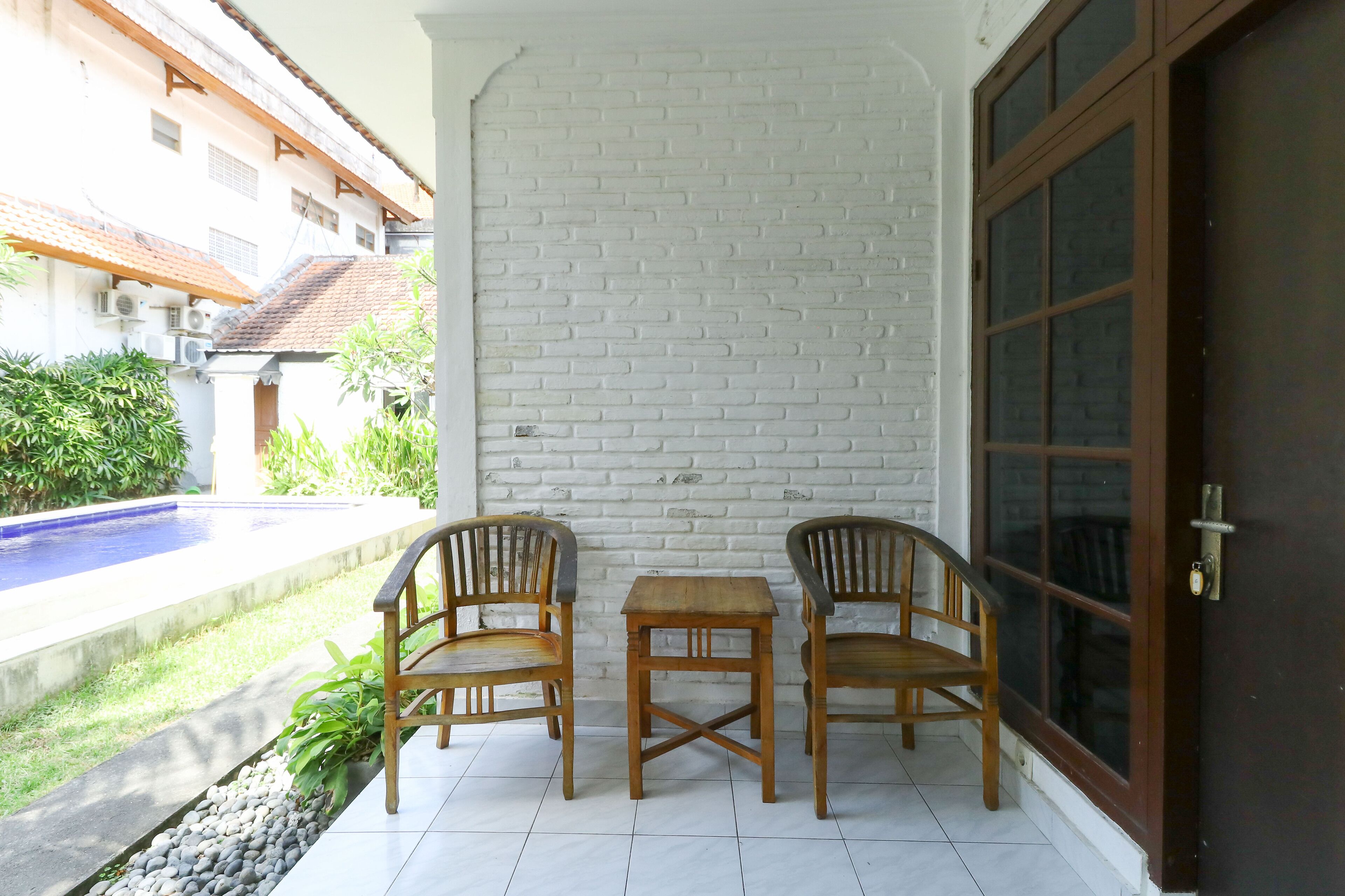 A small patio with two wooden chairs and a table, a white brick wall, and a view of a swimming pool.