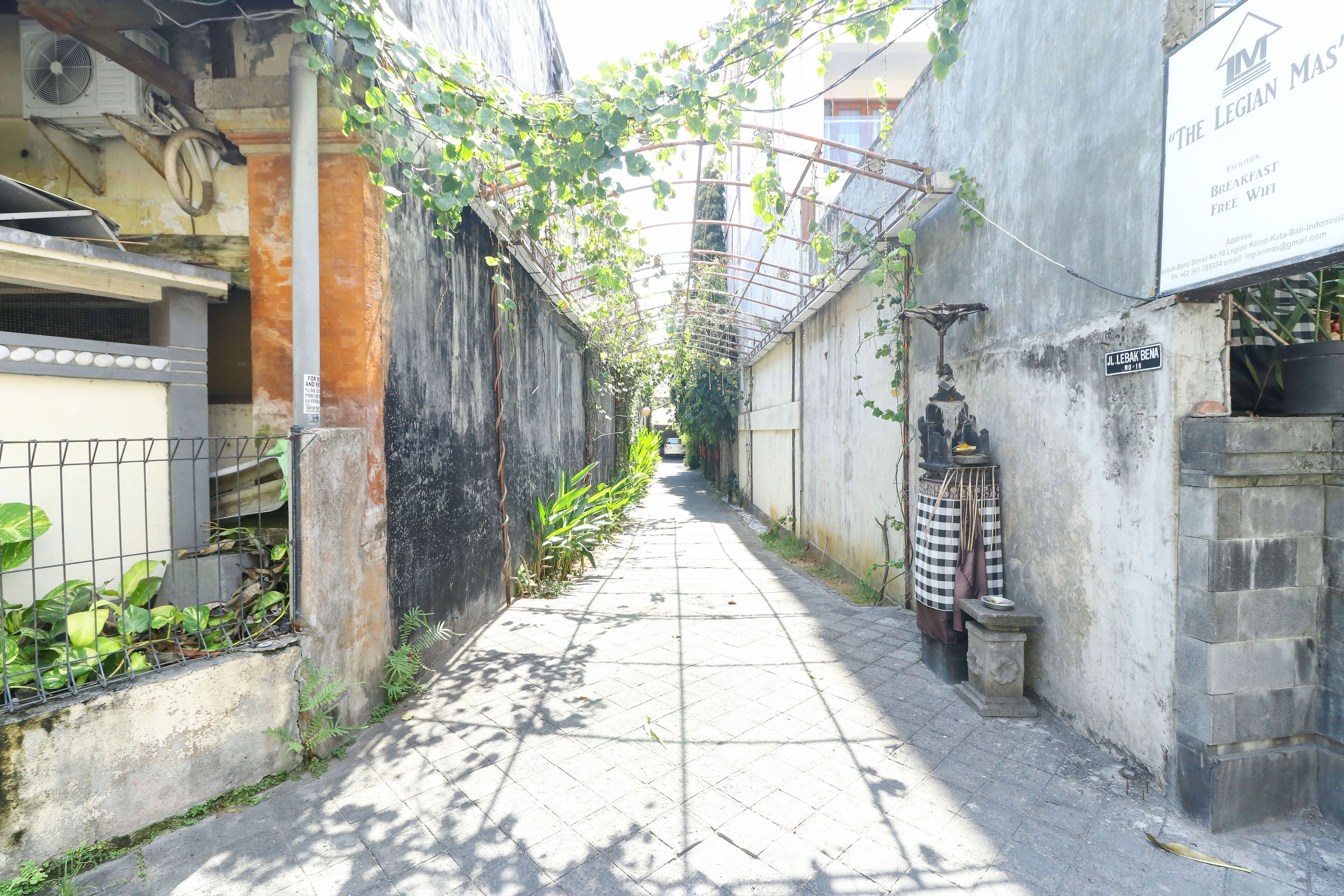 Une ruelle étroite avec un trottoir dallé, une enseigne indiquant « La messe de la Légion », et un petit sanctuaire où sont déposées des offrandes.