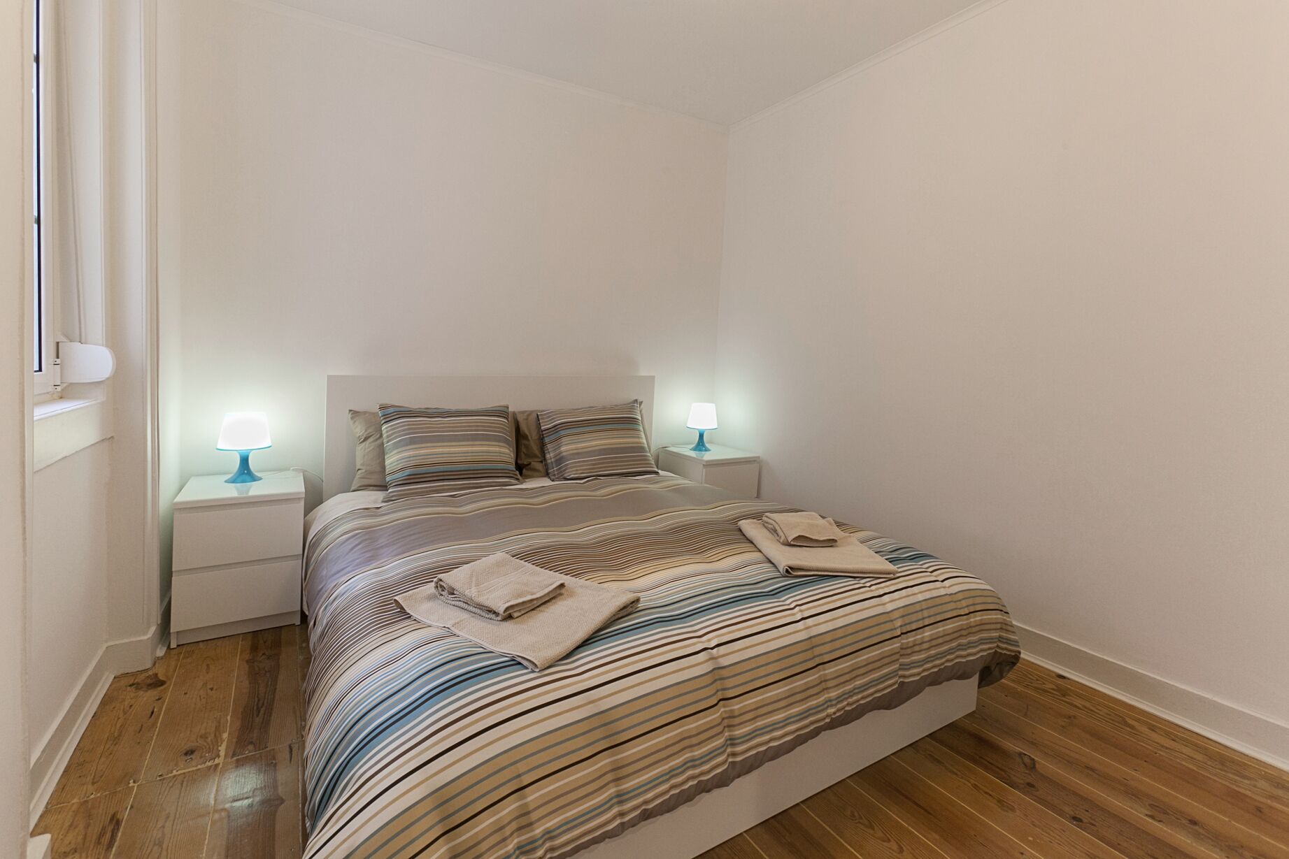 A neatly made bed with striped bedding, two bedside lamps, and a window with blinds.