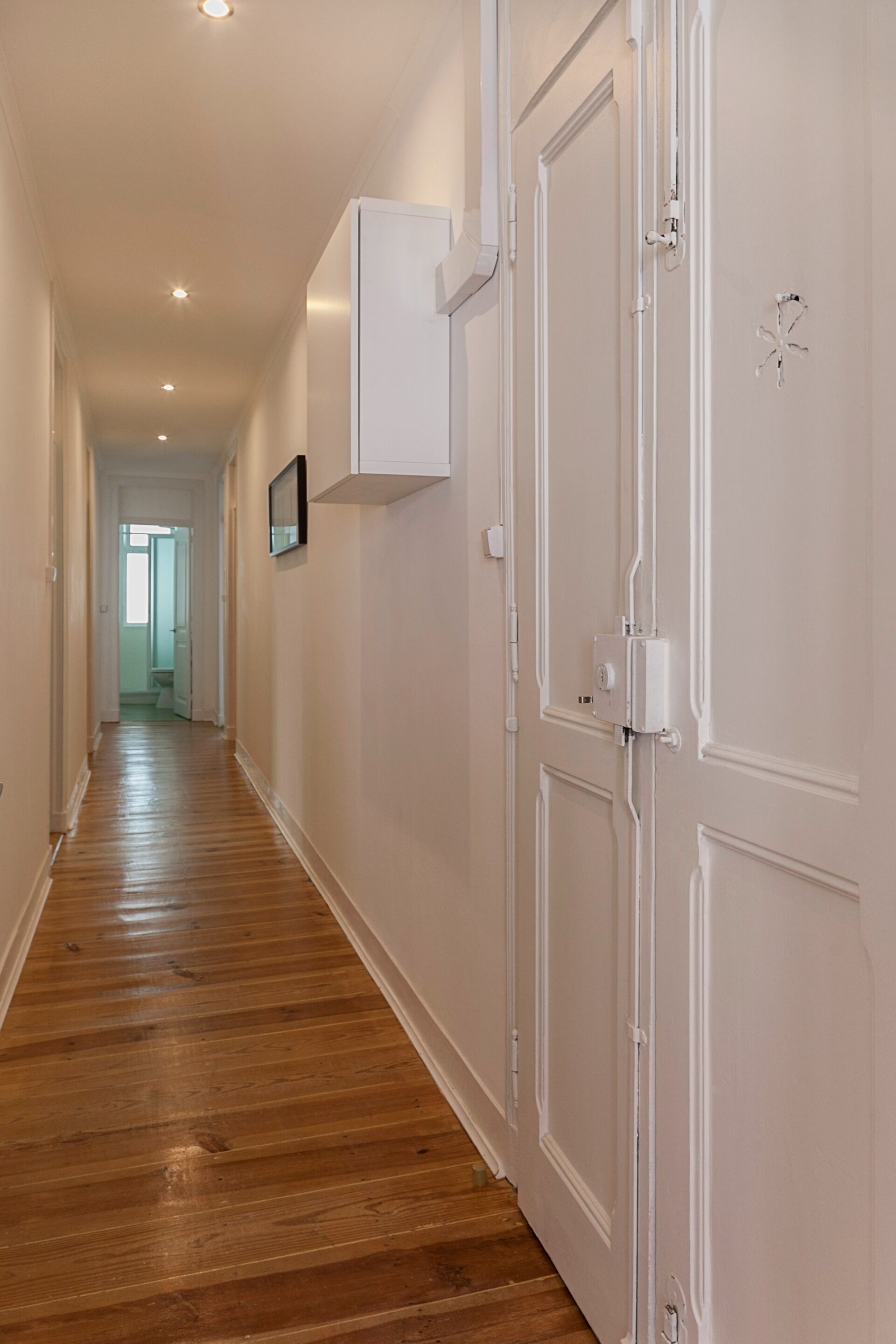 A hallway with wooden flooring, white walls, and a white door with a small electrical box on it.