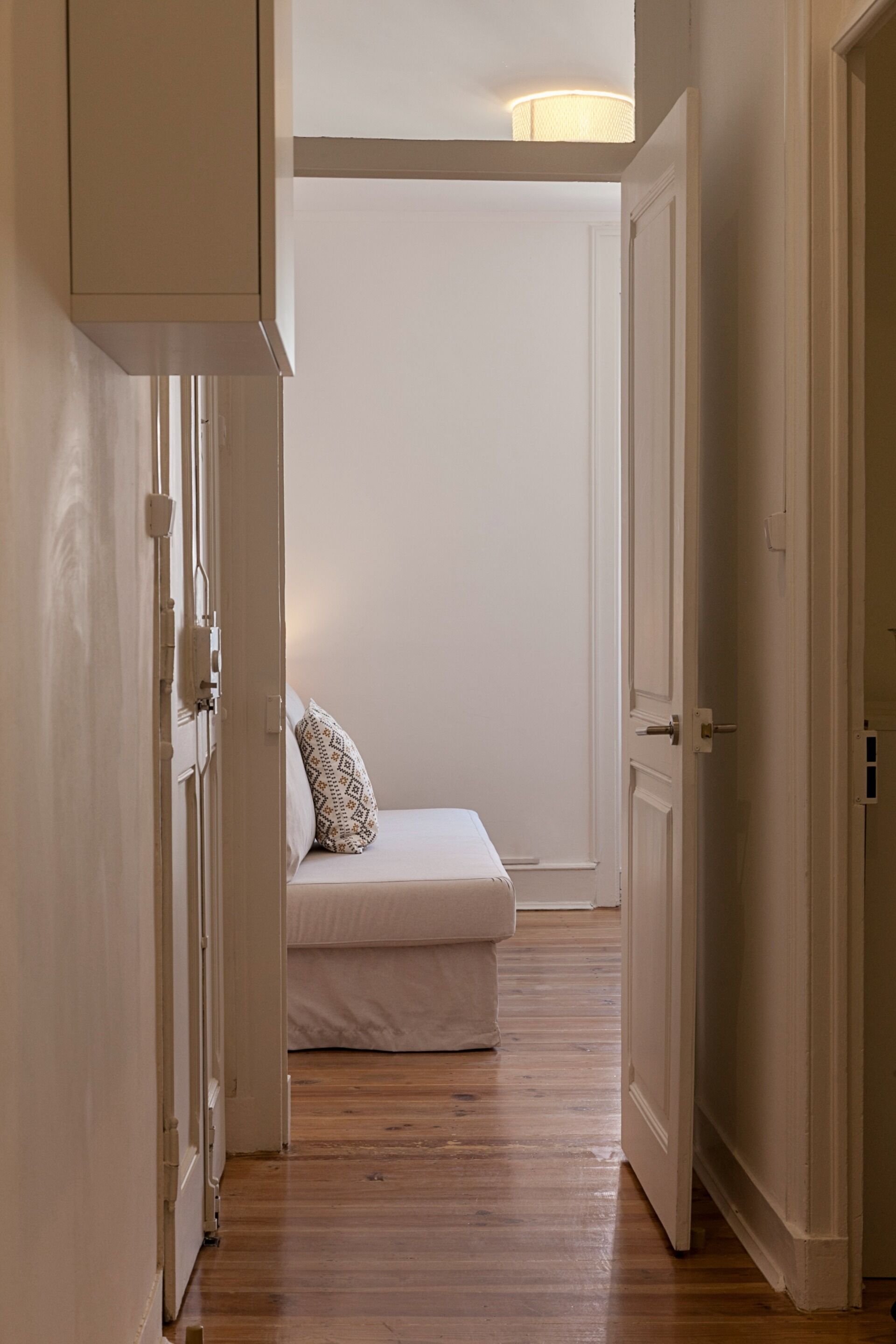 A narrow hallway with wooden flooring leading to a room with a bed and a nightstand.
