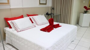 A neatly made bed with white bedding and red pillows, a glass table with a lamp, and a small refrigerator. - Canada Hotel (Rio Verde)