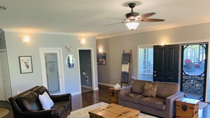 Un salon spacieux avec un canapé en cuir foncé, un canapé de couleur claire et une table basse en bois. On y trouve également un ventilateur de plafond et une porte donnant sur l’extérieur.