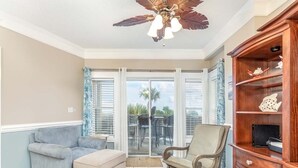 Una habitación con ventilador de techo, un mueble de madera con objetos decorativos, un sillón azul, un sillón beige y un ventanal corredero que da a un área exterior con una mesa y sillas.