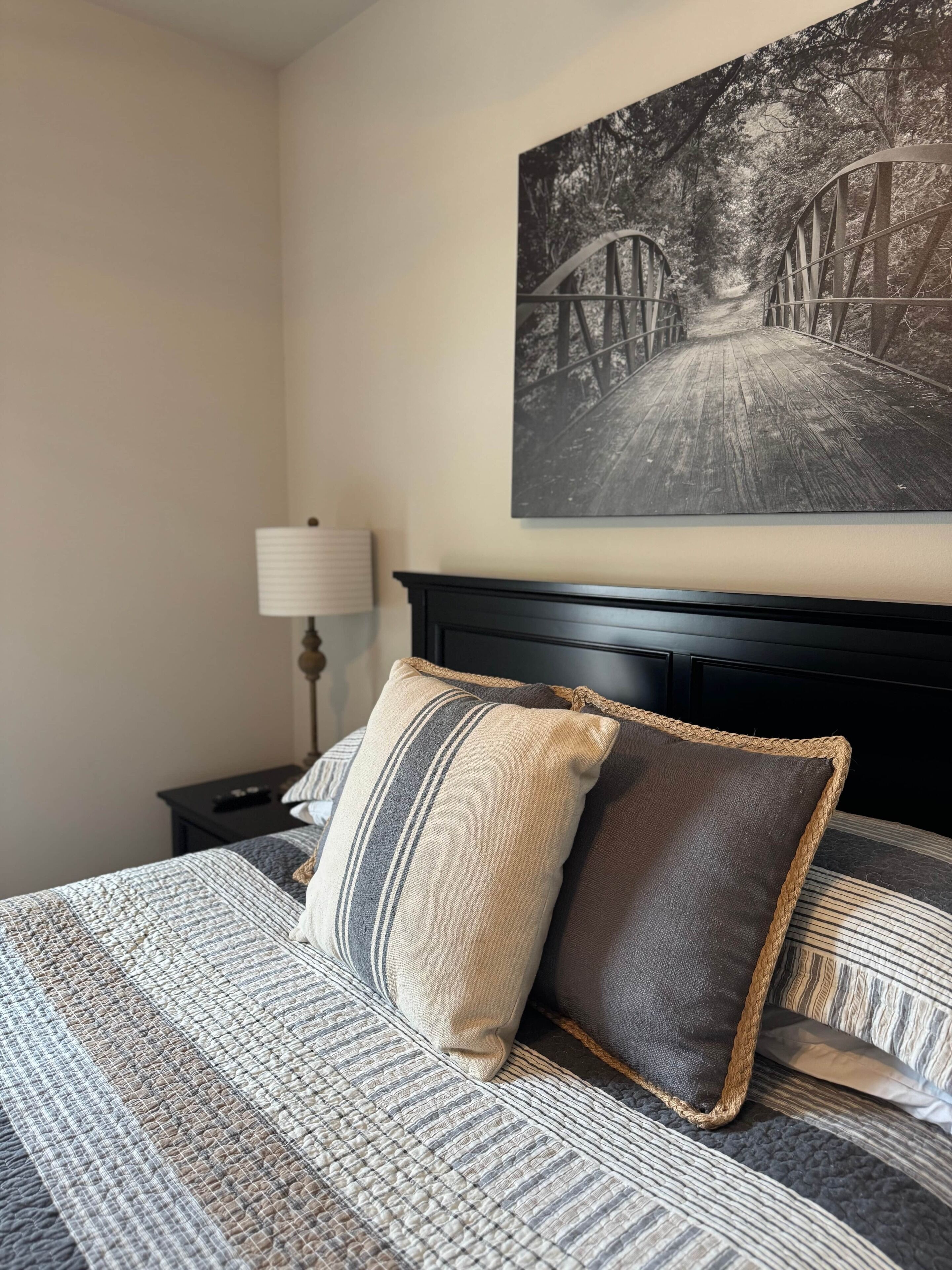 Un lit bien fait, avec une literie à rayures et deux oreillers, une tête de lit noire, une lampe sur une table de chevet et une photographie en noir et blanc d’un pont accrochée au mur.