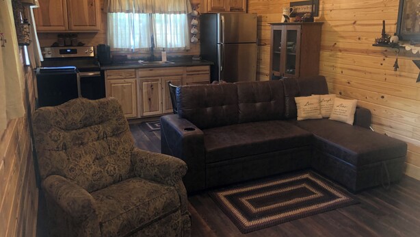 L’intérieur d’un chalet accueillant, avec un salon, un canapé, un fauteuil à motifs, un coin cuisine équipé d’un réfrigérateur, d’une cuisinière et d’une fenêtre agrémentée de rideaux.