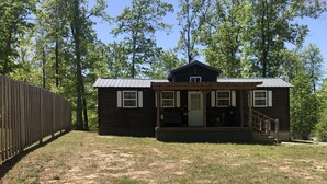 Une petite cabane en bois, de plain-pied, avec une véranda, entourée d’une clôture en bois et d’arbres.