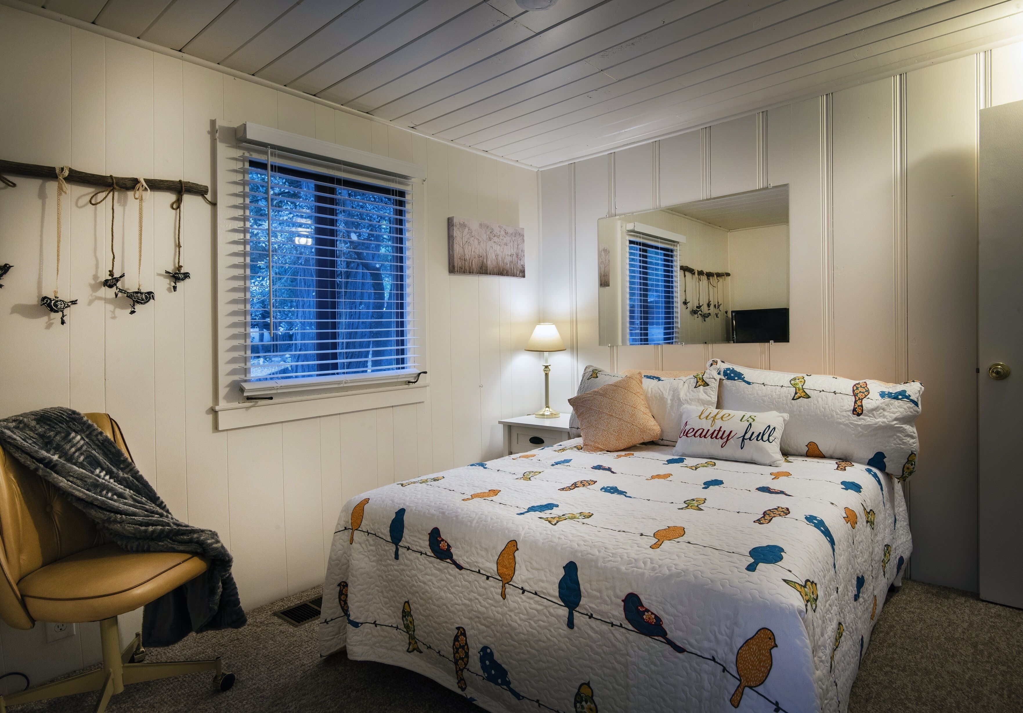 A bedroom with a bed, a chair, a window with blinds, a lamp, a mirror, and wall decorations.
