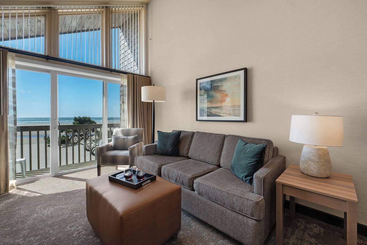 A modern living room with a grey sofa, a brown ottoman, and a side table with a lamp. A framed abstract painting is on the wall, and a chair is positioned near the window.