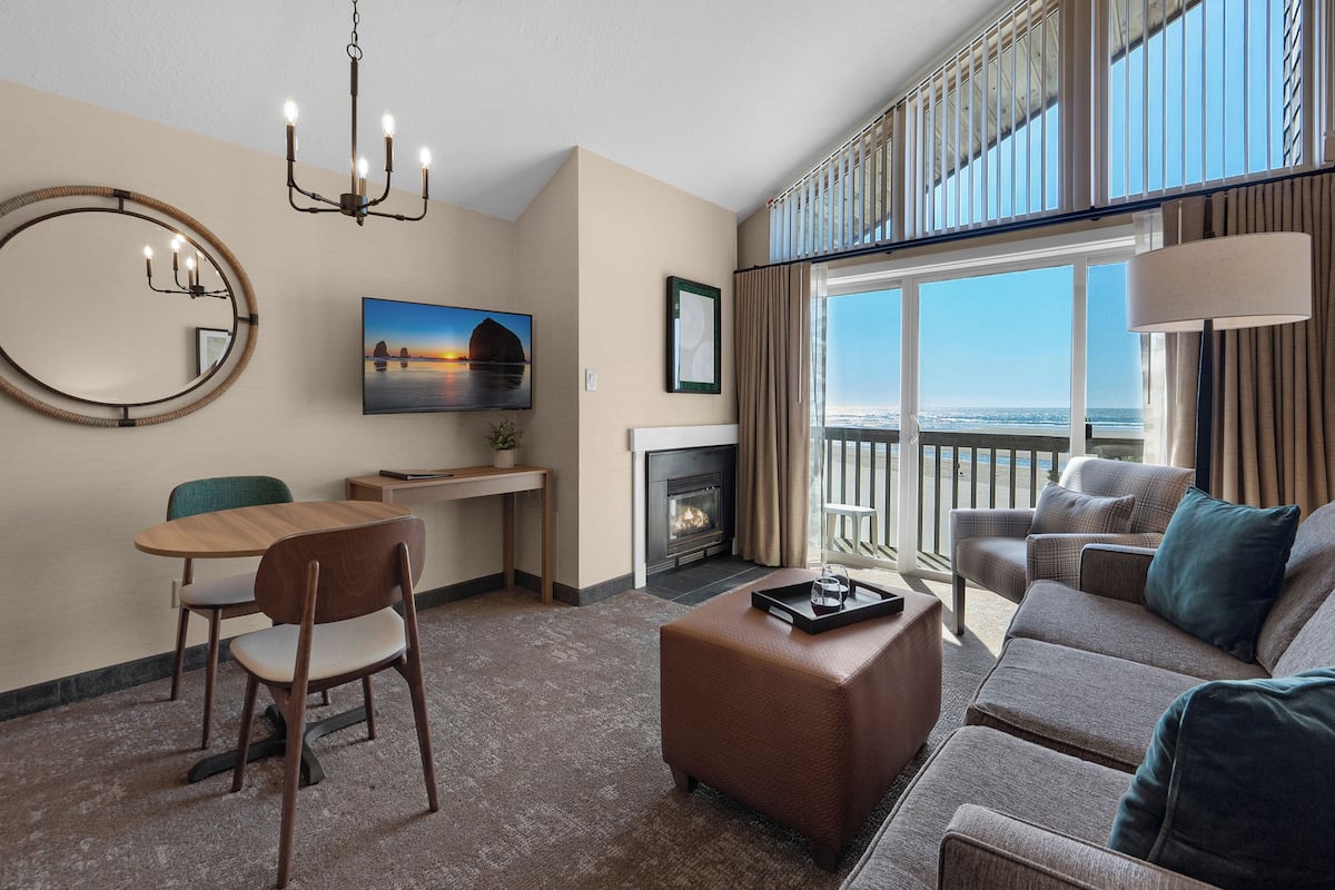A living room with a fireplace, a dining area with a table and chairs, a sofa, and a large window with a view of the ocean.