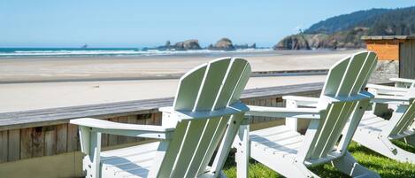 Two Adirondack chairs facing a beach with a wooden deck and a wooden structure in the background.