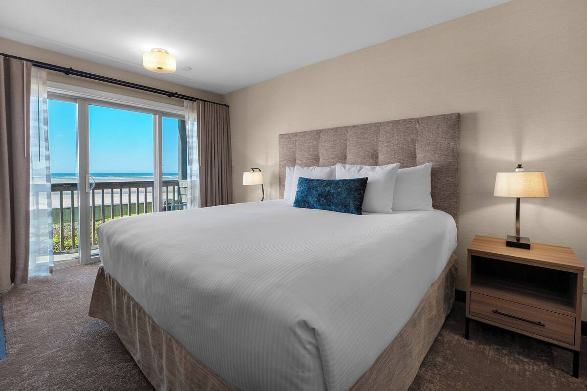 A large bed with a headboard, two bedside lamps, a wooden nightstand, and a view of the ocean through a sliding glass door.