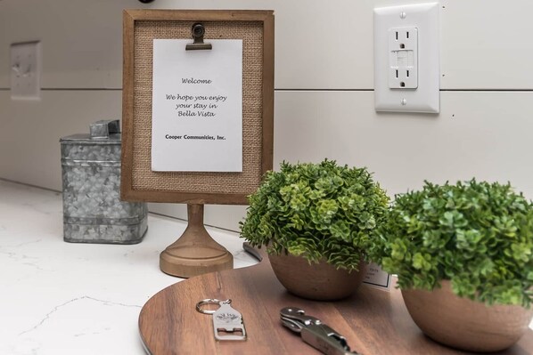 Un letrero de bienvenida, un atril de madera, dos plantas en macetas, un llavero metálico y un recipiente metálico sobre una encimera blanca.