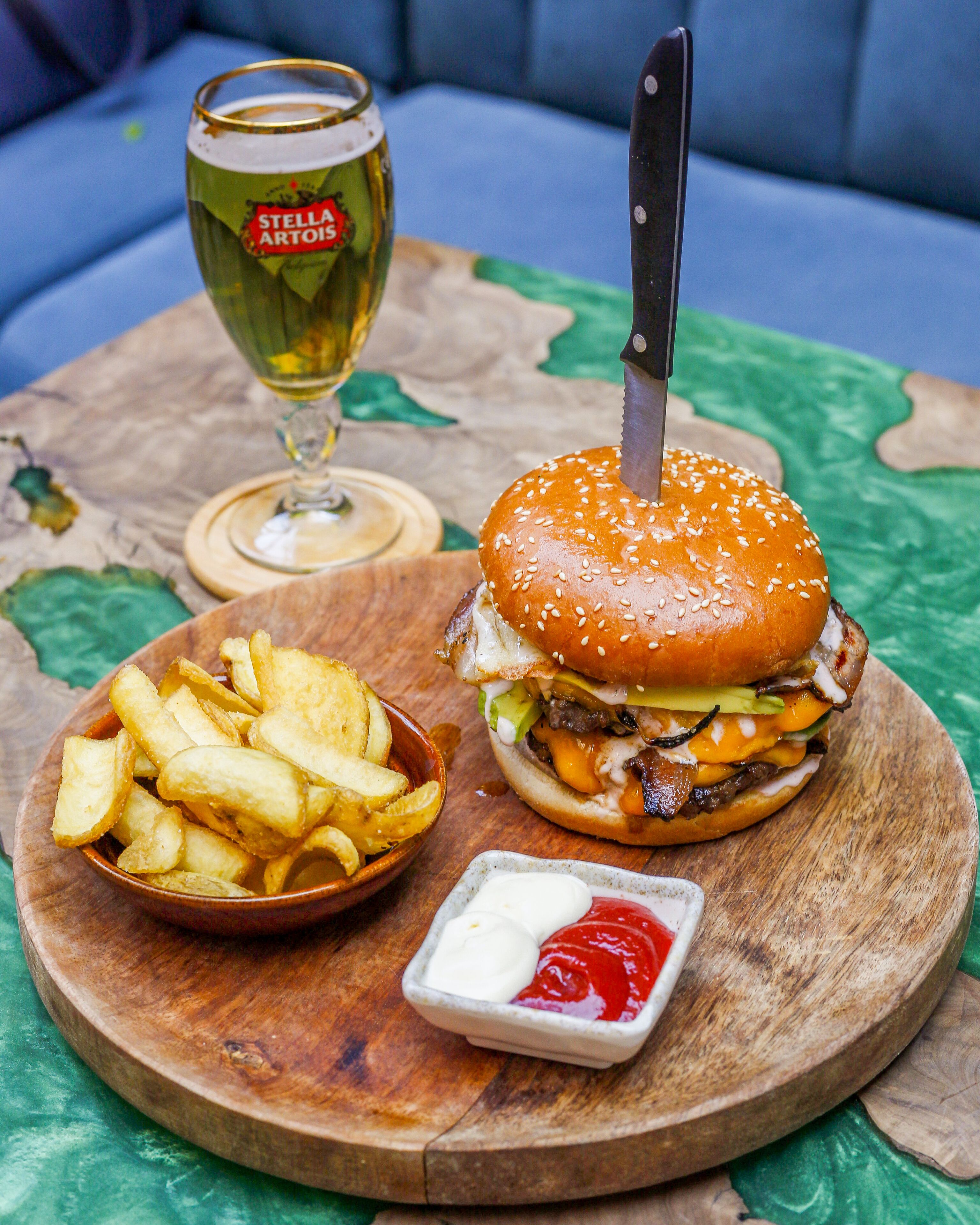 A burger with a knife in it, a glass of beer, a bowl of fries, and a small dish of ketchup and mayonnaise.