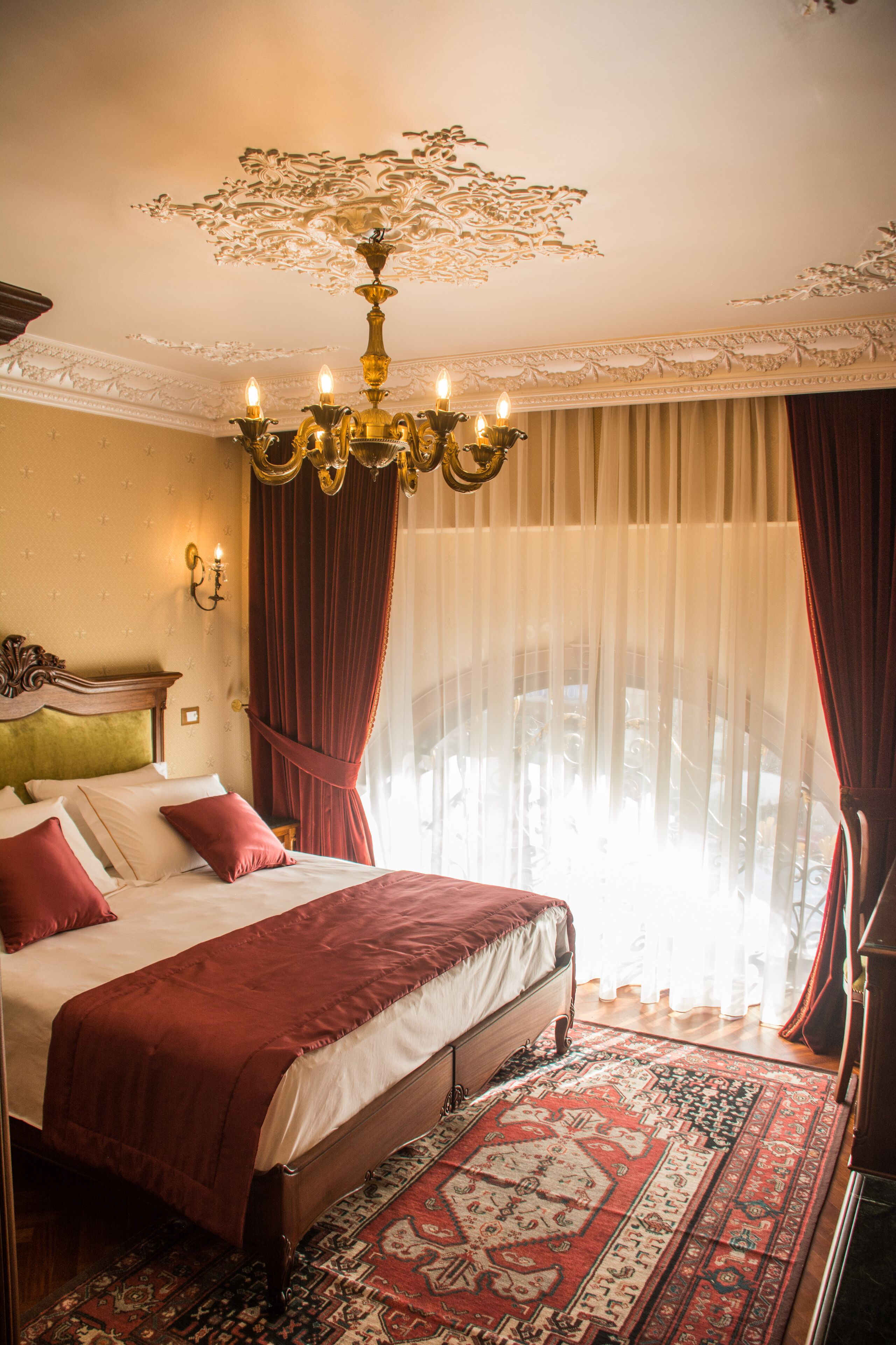 Une chambre d’hôtel avec un grand lit, un lustre somptueux et une vue sur la ville à travers de fines rideaux.
