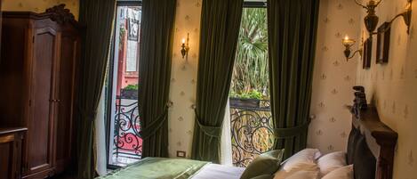 A hotel room with a bed, green curtains, a chandelier, and a view of a balcony with plants.